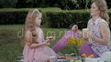一对幸福的母女正在面纱上喝茶.. 一家人在城市公园里，在日落的温暖的晚上野餐。